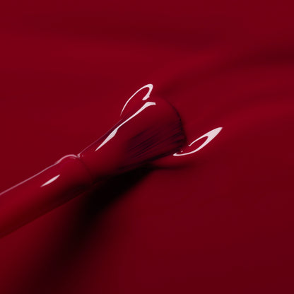 A close-up of the Manucurist Violeta nail polish brush coating a surface with glossy burgundy purple polish. The bristles and handle are partly covered in the shiny polish, creating a sleek, vibrant look.