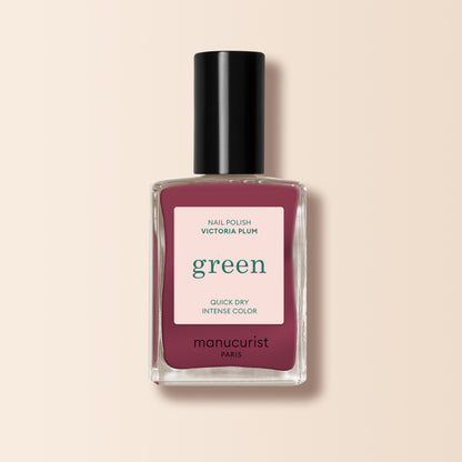 A bottle of Manucurist Victoria Plum nail polish, featuring a deep plum shade, black cap, and white label with green and pink text, is displayed on a light beige background.