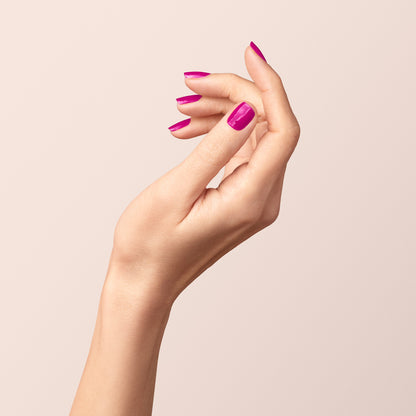 A hand with light skin is raised against a plain light background, nails polished in bright pink "Armeria" gel by Manucurist, with gently curved fingers in a relaxed pose.