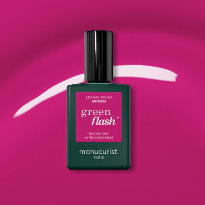 A bottle of Manucurist Armeria rests on a vibrant pink background with a curved mauve streak behind it. The dark green bottle features a black cap and a bright pink label.