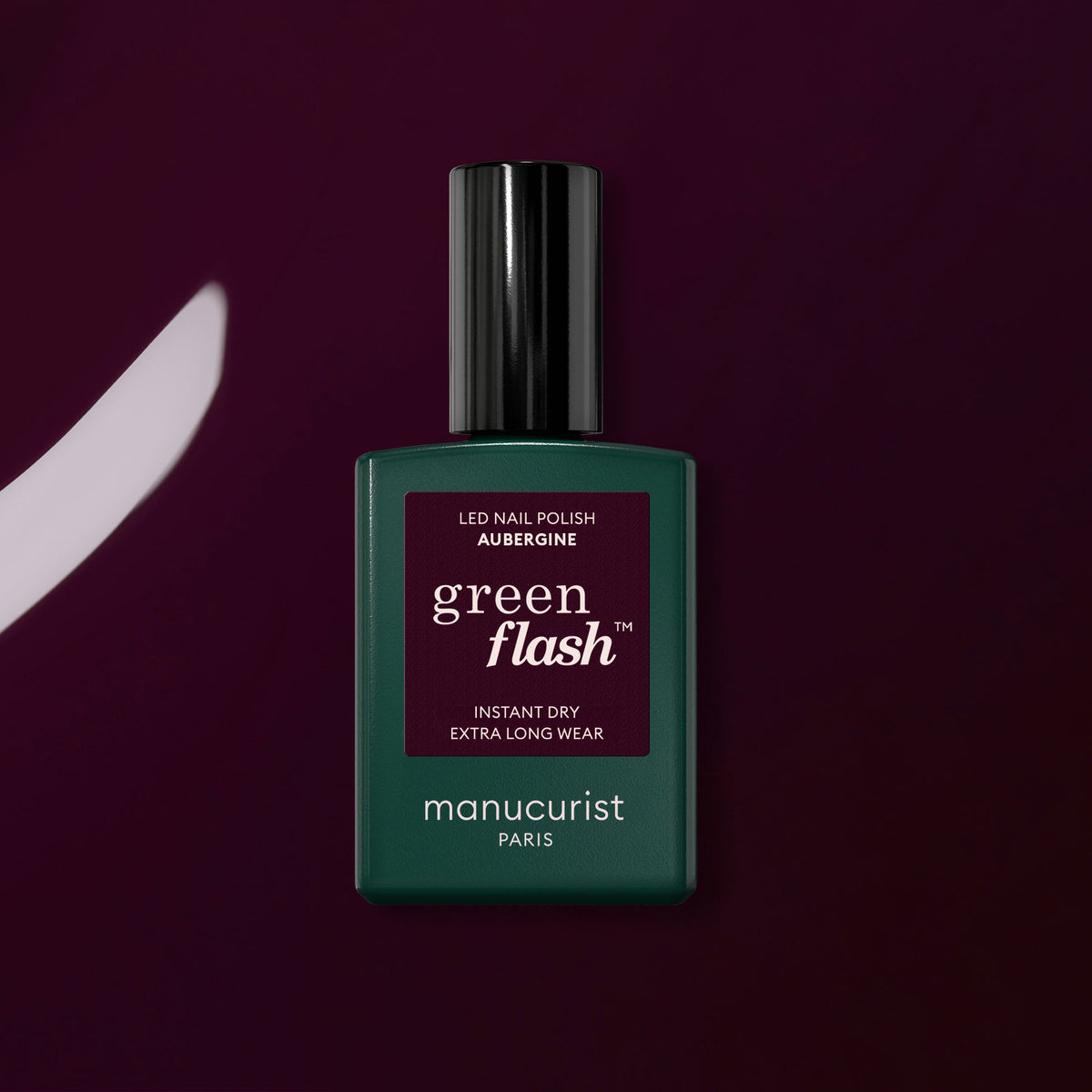 A bottle of Manucurist Aubergine Green Flash gel polish stands upright on a deep purple surface, with a matching brushstroke displayed to its left.