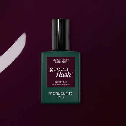 A bottle of Manucurist Aubergine Green Flash gel polish stands upright on a deep purple surface, with a matching brushstroke displayed to its left.