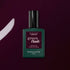 A bottle of Manucurist Aubergine Green Flash gel polish stands upright on a deep purple surface, with a matching brushstroke displayed to its left.