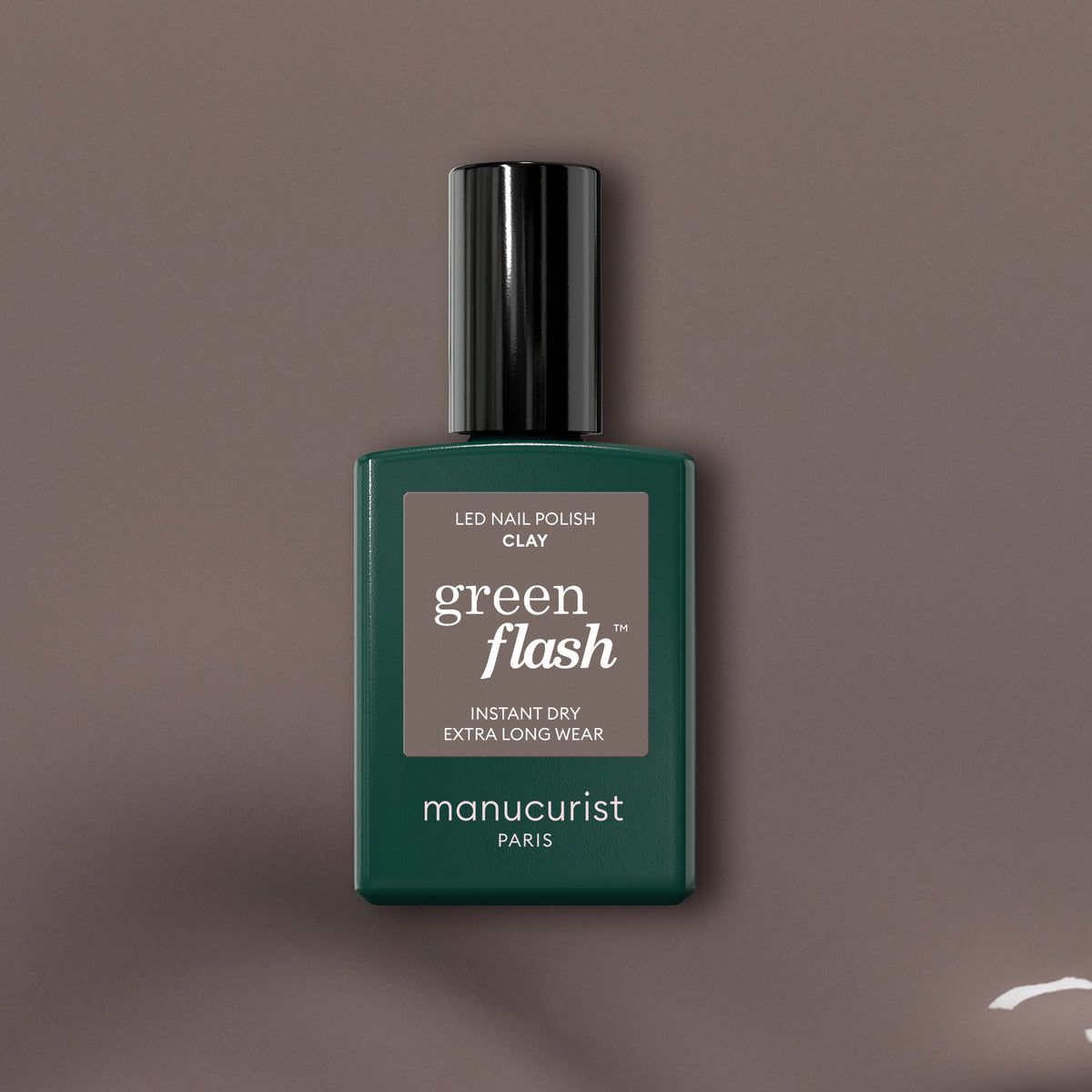 Un flacon de vernis gel Green Flash Clay de Manucurist est centré sur un fond taupe, mettant en valeur le flacon vert foncé avec son bouchon noir et son texte blanc qui souligne cette teinte taupe innovante.