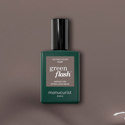 Un flacon de vernis gel Green Flash Clay de Manucurist est centré sur un fond taupe, mettant en valeur le flacon vert foncé avec son bouchon noir et son texte blanc qui souligne cette teinte taupe innovante.