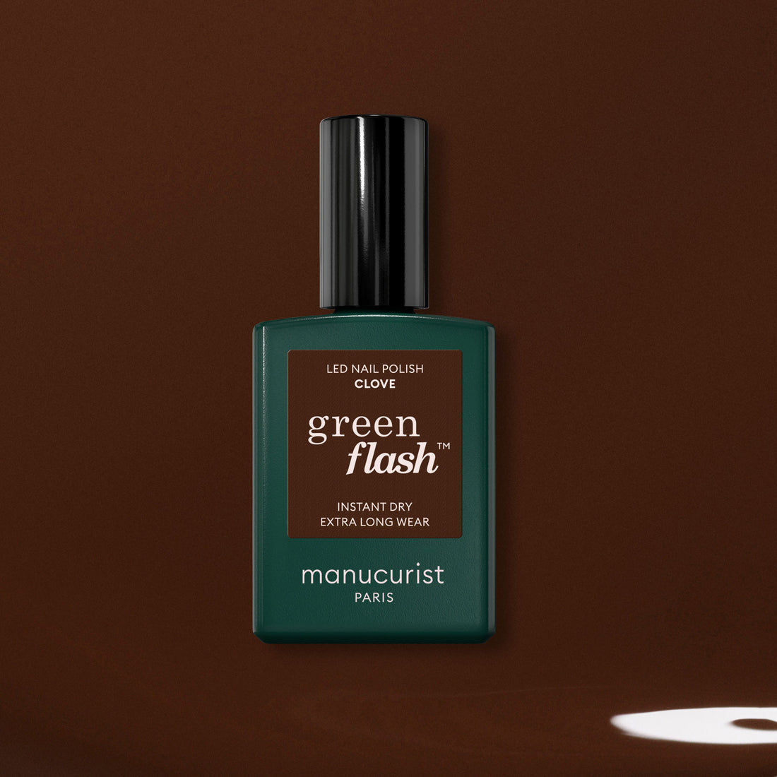 Un flacon de vernis gel Manucurist Clove, une teinte brun chocolat intense, est présenté sur un fond marron avec une petite tache claire près du coin inférieur droit.