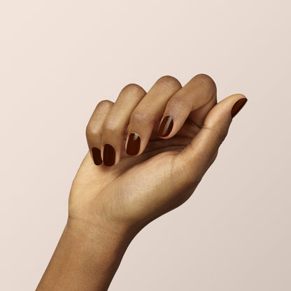 A hand with medium brown skin is shown against a pale background. The nails are polished in Manucurist’s Clove, a glossy chocolate brown shade, with one nail featuring a nude geometric accent.