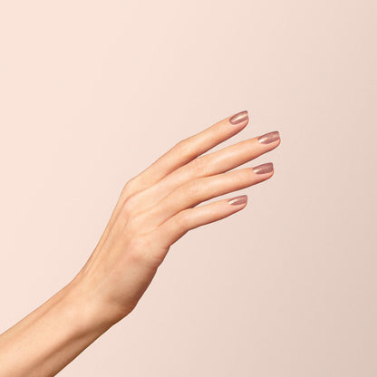 Gros plan sur une main aux doigts fins et aux ongles soigneusement manucurés, peints avec Cosmic Rose de Manucurist, un vernis rose irisé, posée avec grâce sur un fond beige doux.