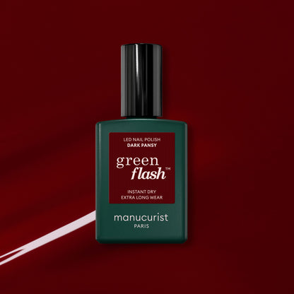 A bottle of Manucurist Dark Pansy gel polish rests on a glossy, deep red background with a matching swipe of velvet red nail color beside it.