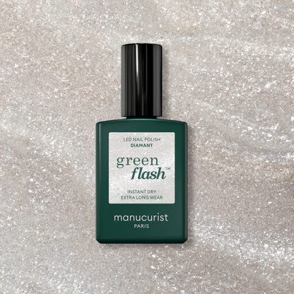A bottle of Manucurist Diamant gel polish rests on a sparkly, light surface. The dark green bottle with a black cap and white label showcases the shimmering silver nail color.