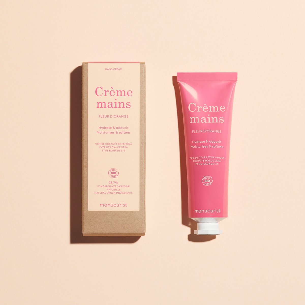 Un tube rose de crème pour les mains Manucurist Everyday Orange Blossom - Cosmos Organic est posé à côté de sa boîte rectangulaire marron sur un fond beige clair, les deux articles affichant du texte en français.