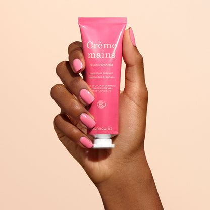 Une main avec du vernis à ongles rose tient un tube rose de crème pour les mains Manucurist Everyday Orange Blossom - Cosmos Organic, enrichie en huile de coco, sur un fond beige.