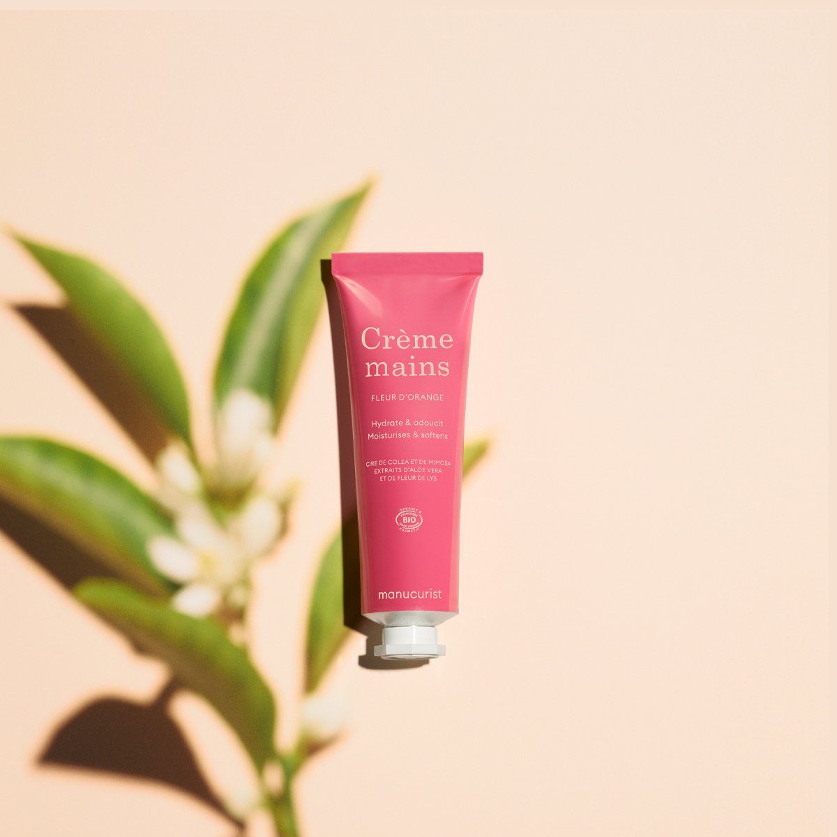 Un tube rose de crème pour les mains Manucurist Everyday Orange Blossom Hand Cream - Cosmos Organic se tient debout sur un fond beige, avec du texte blanc et de l’huile de coco, tandis qu’une plante feuillue projette une ombre douce à proximité.