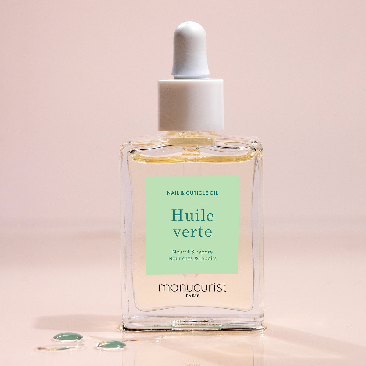 A glass dropper bottle of Manucurist Nourishing Cuticle Oil sits on a light surface with a few oil drops nearby. The pale green label with white and dark green text suggests enhanced nail hydration.