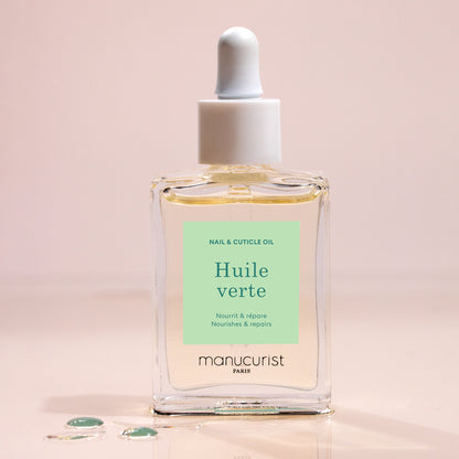 A glass dropper bottle of Manucurist Nourishing Cuticle Oil sits on a light surface with a few oil drops nearby. The pale green label with white and dark green text suggests enhanced nail hydration.