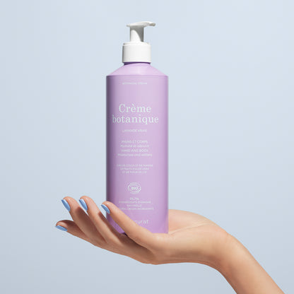Une main aux ongles vernis tient un flacon pompe lavande intitulé Soothing Lavender Hand  Body Cream - Cosmos Organic by Manucurist, un soin hydratant bio pour les mains et le corps, sur fond bleu clair.