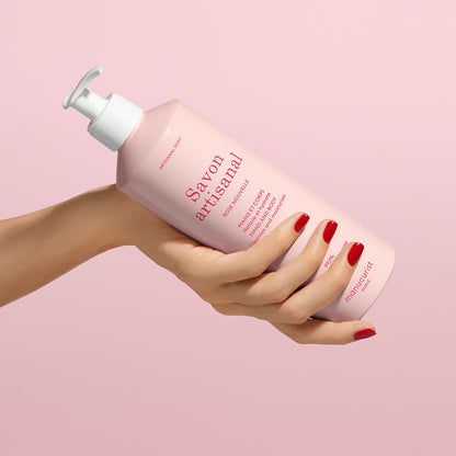 A hand with red nail polish holds a pink bottle of Manucurist Organic Rose Hand  Body Wash - Cosmos Organic with a white pump, set against a matching pink background.