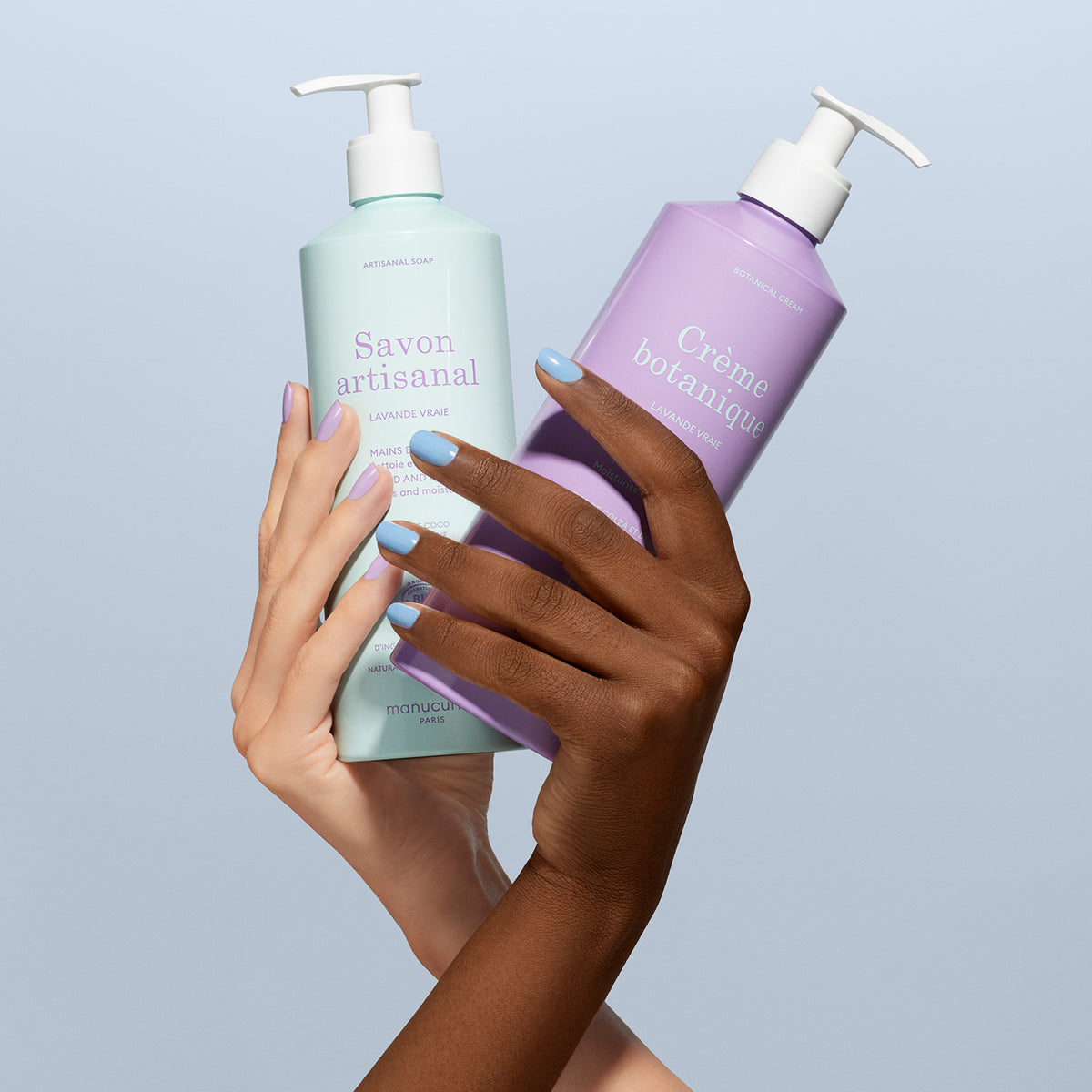 Two hands, one light and one dark-skinned, with matching pastel blue nails, hold Manucurist’s Lavender Botanical Cream  Artisan Soap Duo - Cosmos Organic against a light blue background.