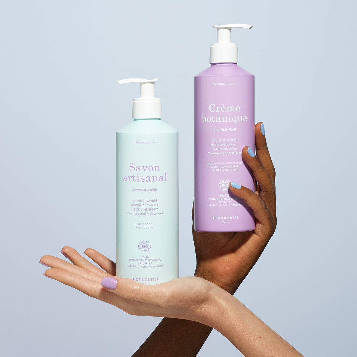 Two hands, one light and one dark-skinned, hold the Manucurist Lavender Botanical Cream  Artisan Soap Duo - Cosmos Organic against a pale blue background. The cream is infused with real lavender and the soap is mint green.