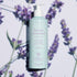 A light blue bottle of Manucurist Organic Lavender Hand  Body Wash - Cosmos Organic with a pump sits on a surface with lavender flowers and soft shadows. The label highlights coconut and olive oil, natural ingredients, and a subtle lavender scent.