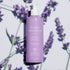 Un flacon violet de crème pour les mains et le corps Soothing Lavender de Manucurist - Cosmos Organic, avec une pompe blanche, est posé sur une surface blanche, encadré de fleurs de lavande et d’ombres. L’étiquette française met en avant son essence botanique et sa formule hydratante.
