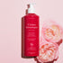 A pink bottle of Manucurist Soothing Rose Hand  Body Cream - Cosmos Organic stands upright on a light pink background beside two pink roses. The white pump and text highlight its promise to deeply hydrate your skin.