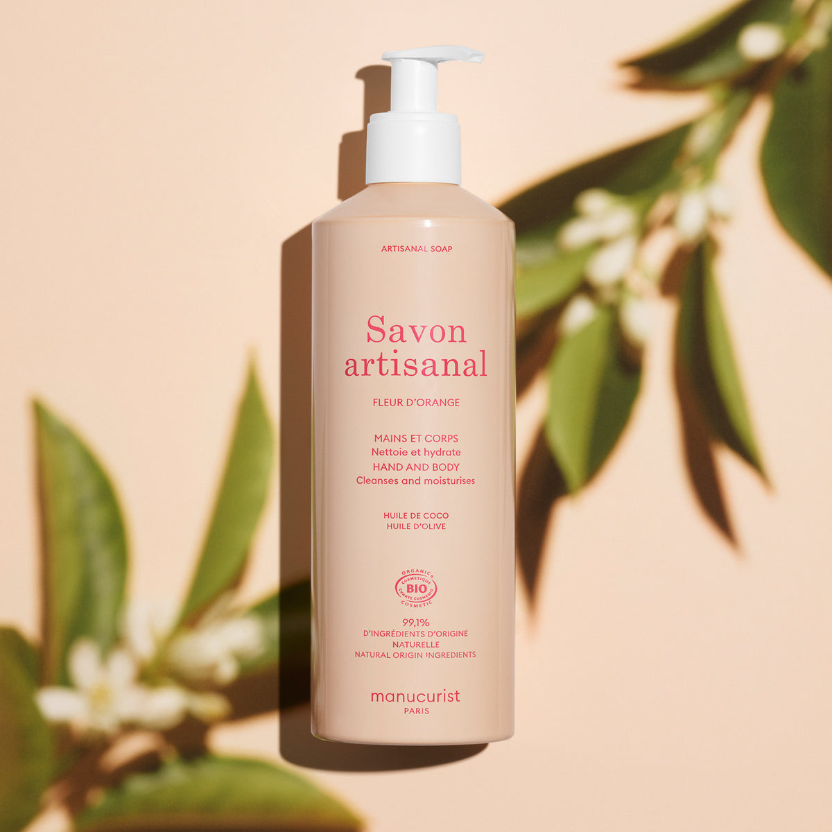 A beige pump bottle of Manucurist Organic Orange Blossom Hand  Body Wash - Cosmos Organic stands upright on a peach background, surrounded by green leaves and their shadows, showcasing this artisan soap infused with orange blossom.