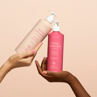 Two hands hold up Manucurist’s Orange Blossom Botanical Cream  Artisan Soap Duo - Cosmos Organic against a light peach background: the left holds the artisan soap, while the right displays the bright pink botanical cream.