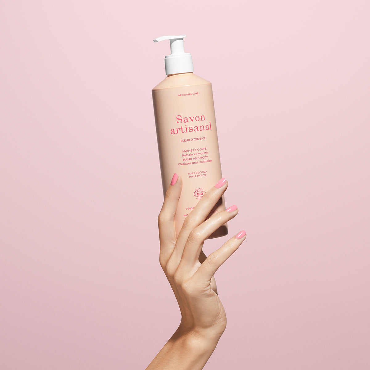 A manicured hand holds a pink bottle of Manucurist Organic Orange Blossom Hand  Body Wash – Cosmos Organic, set against a pale pink background.
