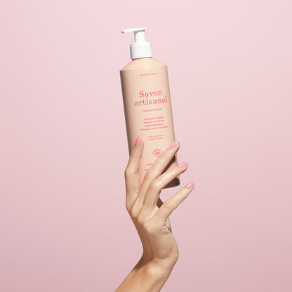 A manicured hand holds a pink bottle of Manucurist Organic Orange Blossom Hand  Body Wash – Cosmos Organic, set against a pale pink background.