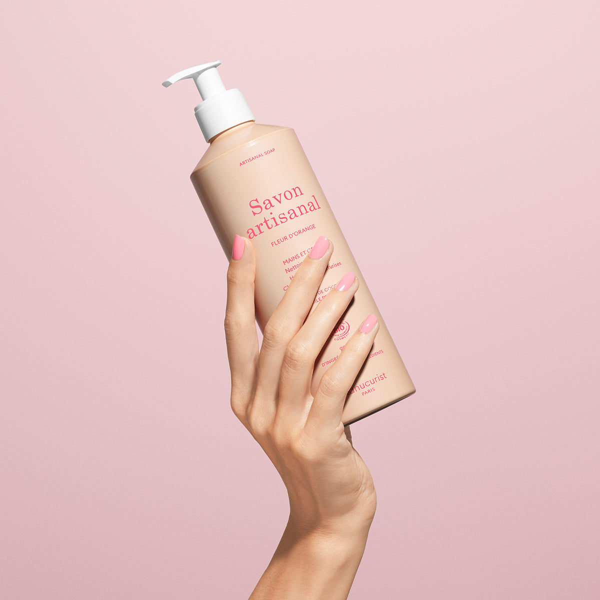 A hand with manicured pink nails holds up a bottle of Manucurist Organic Orange Blossom Hand  Body Wash - Cosmos Organic with a pump dispenser against a pale pink background.