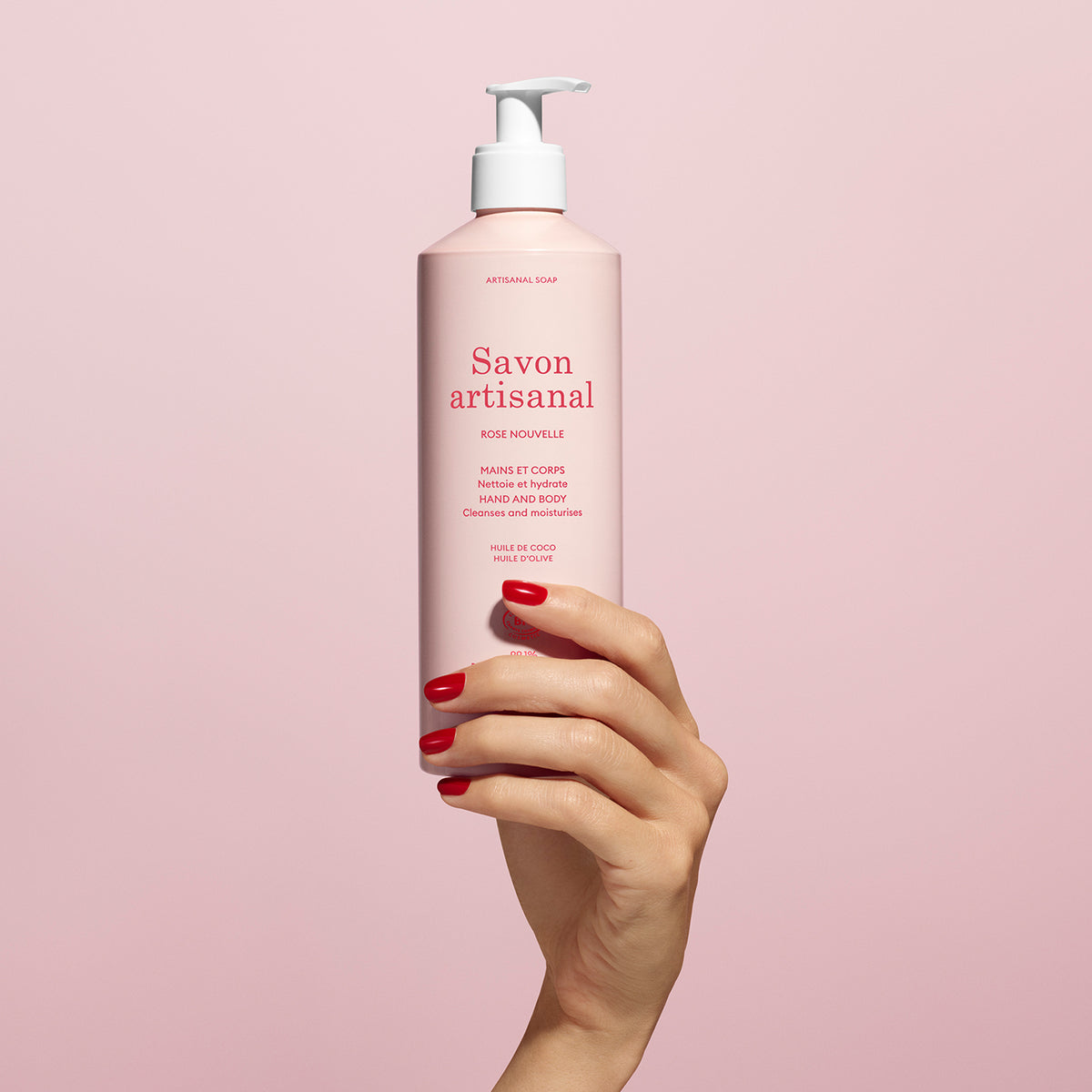 A hand with red-painted nails holds a pink bottle of Manucurist Organic Rose Hand  Body Wash - Cosmos Organic with a white pump and red text, set against a pale pink background.