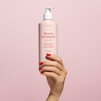 A hand with red-painted nails holds a pink bottle of Manucurist Organic Rose Hand  Body Wash - Cosmos Organic with a white pump and red text, set against a pale pink background.