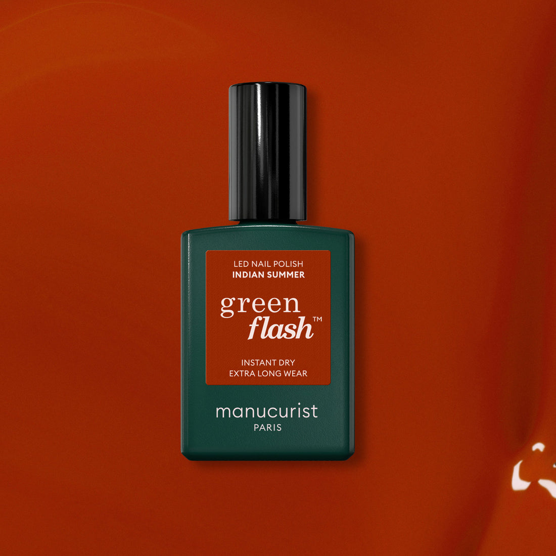 A bottle of Manucurist Indian Summer Green Flash LED nail polish—an ochre red shade—stands against a matching orange-red background, with a dark green bottle, black cap, and white text.