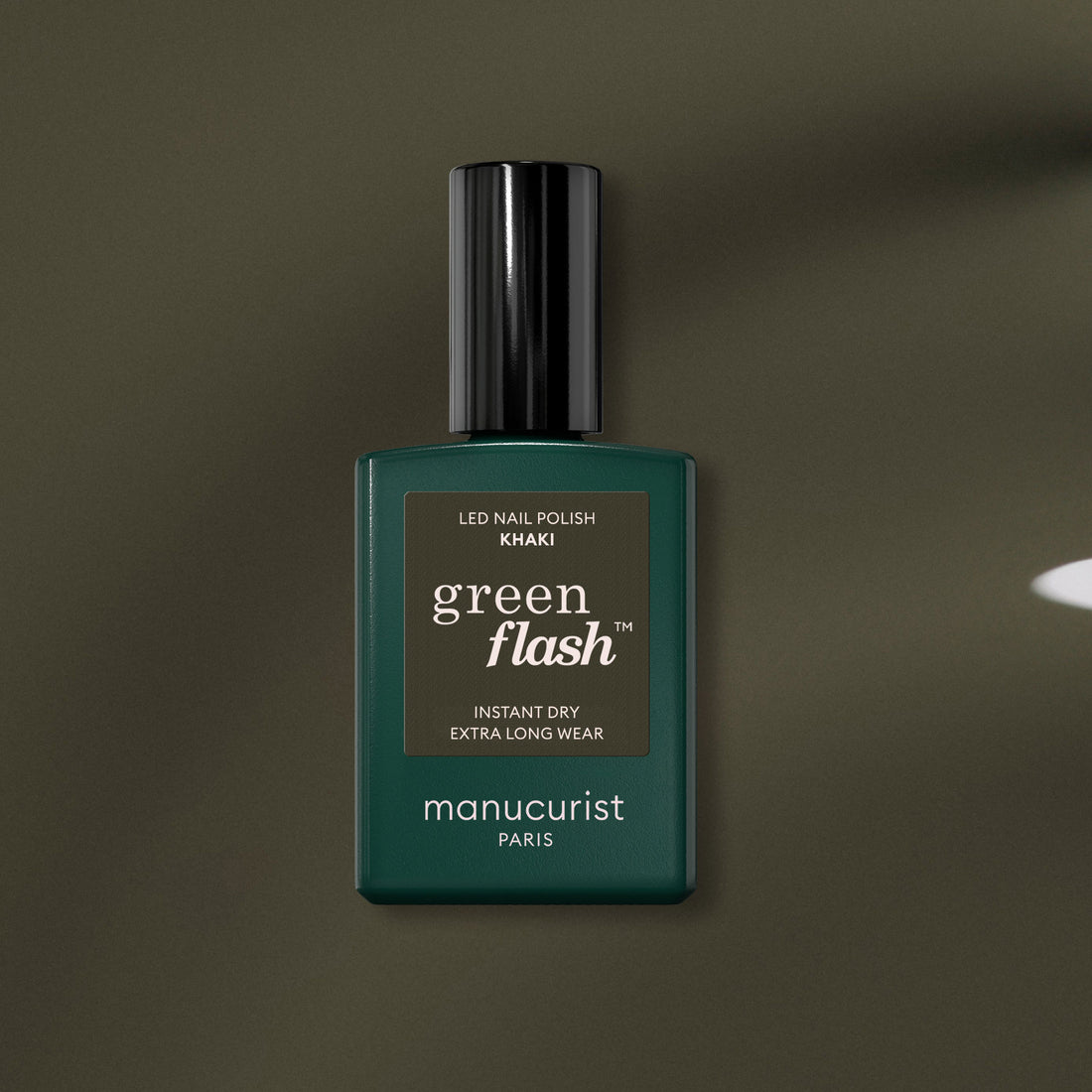 Un flacon de vernis gel Manucurist Khaki aux sous-tons gris, avec une étiquette vert foncé et un bouchon noir, repose sur une surface vert olive.