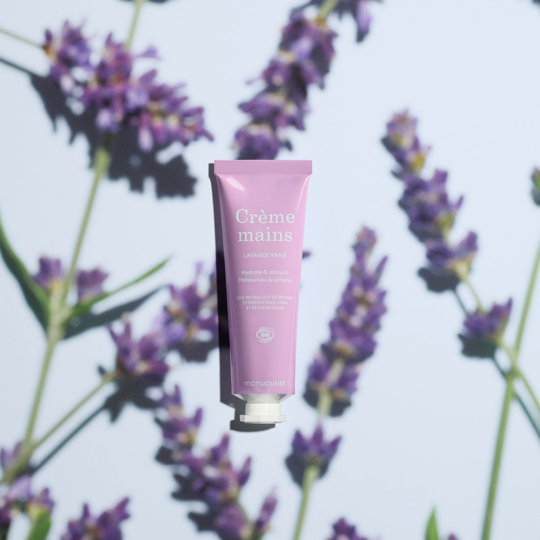 A pink tube of Manucurist Everyday Lavender Hand Cream - Cosmos Organic sits centered on a light background with lavender sprigs, casting soft shadows. This organic hand cream delivers a fresh, soothing lavender scent.
