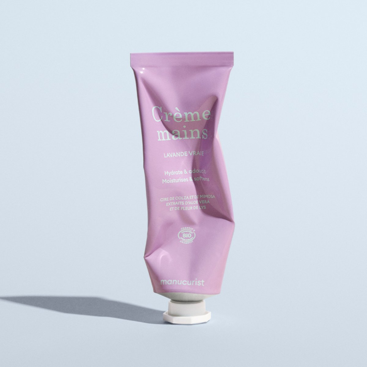 Un tube rose froissé de crème mains Manucurist Everyday Lavender Hand Cream - Cosmos Organic est posé à la verticale sur une surface gris clair en projetant une ombre. L&
