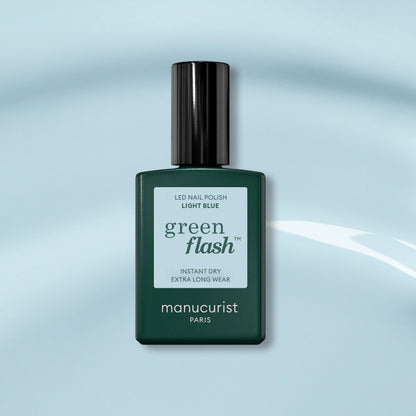 A bottle of Manucurist Light Blue Green Flash gel polish, with a dark green cap, sits on a soft blue background. The label highlights instant dry and extra long wear for this pastel sky blue shade.