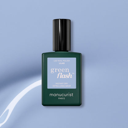 A bottle of Manucurist Lilas Green Flash gel polish, featuring a dark green base, black cap, and light purple label, is set against a light blue background with a white curved streak.