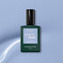 A bottle of Manucurist Lilas Green Flash gel polish, featuring a dark green base, black cap, and light purple label, is set against a light blue background with a white curved streak.