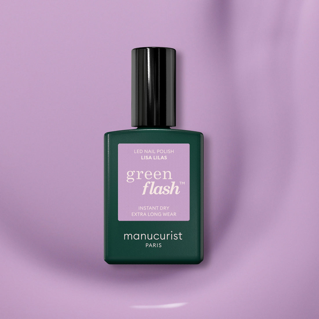 A bottle of Manucurist Lisa Lilas gel polish featuring a dark green base, black cap, and purple label rests on a smooth light purple surface, exuding subtle 90s vibes.