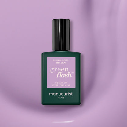 Un flacon de vernis gel Manucurist Lisa Lilas avec une base vert foncé, un bouchon noir et une étiquette violette repose sur une surface lisse de couleur mauve clair, dégageant une subtile ambiance années 90.
