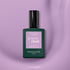 Un flacon de vernis gel Manucurist Lisa Lilas avec une base vert foncé, un bouchon noir et une étiquette violette repose sur une surface lisse de couleur mauve clair, dégageant une subtile ambiance années 90.