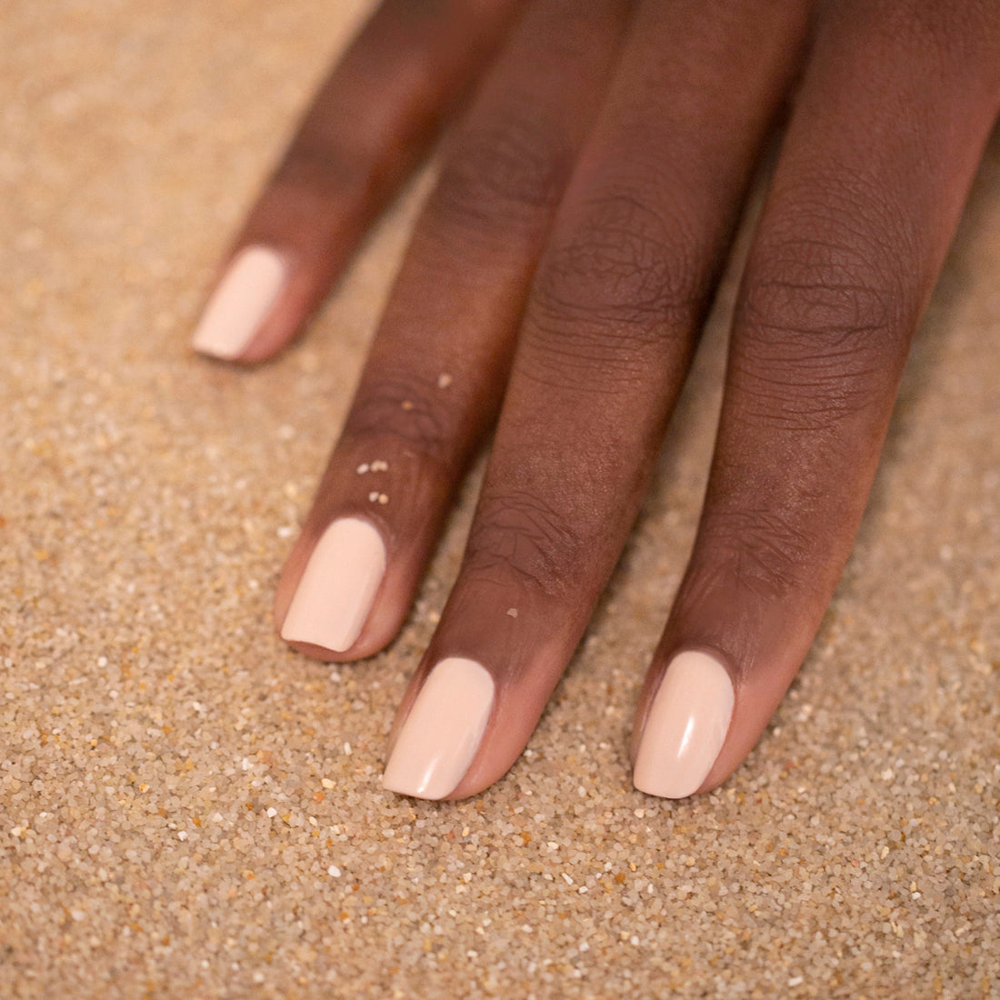 A hand with medium-dark skin tone rests on sand, featuring neatly manicured nails painted in Manucurist&