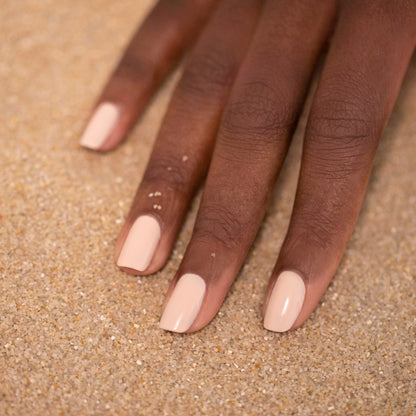 A hand with medium-dark skin tone rests on sand, featuring neatly manicured nails painted in Manucurist&