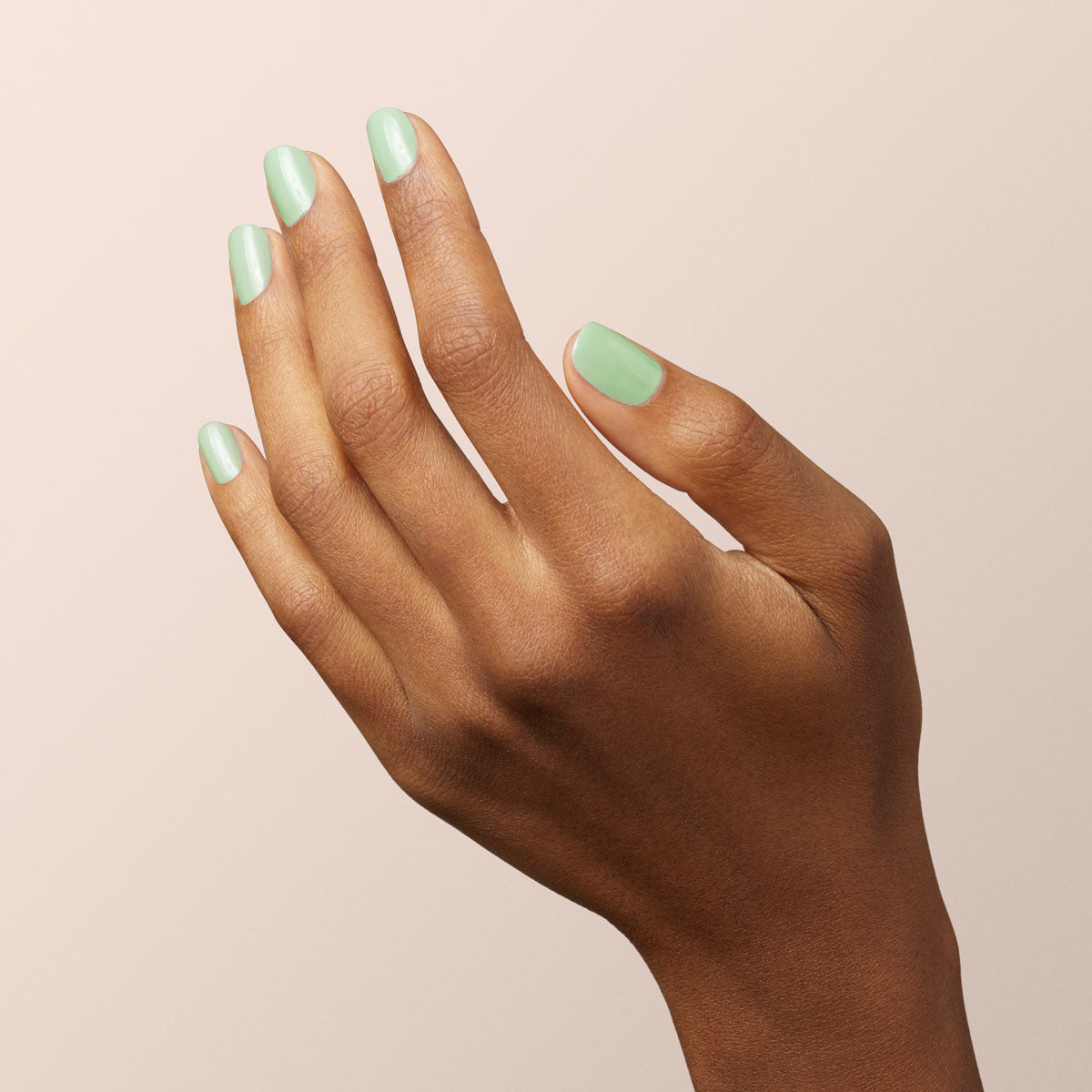 A hand with medium brown skin appears against a light beige background. The manicured nails are painted with "Mint" pastel green nail polish from Manucurist.