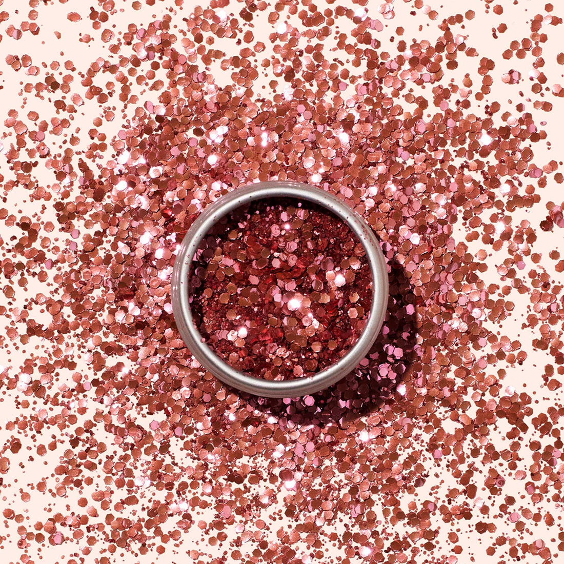 A round metal tin of Manucurist Pink Quartz Biodegradable Glitter, featuring shiny pink hexagonal pieces for nail art, sits amid scattered glitter on a light pink background.