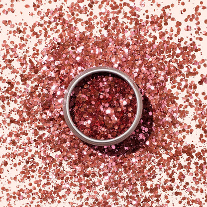 A round metal tin of Manucurist Pink Quartz Biodegradable Glitter, featuring shiny pink hexagonal pieces for nail art, sits amid scattered glitter on a light pink background.