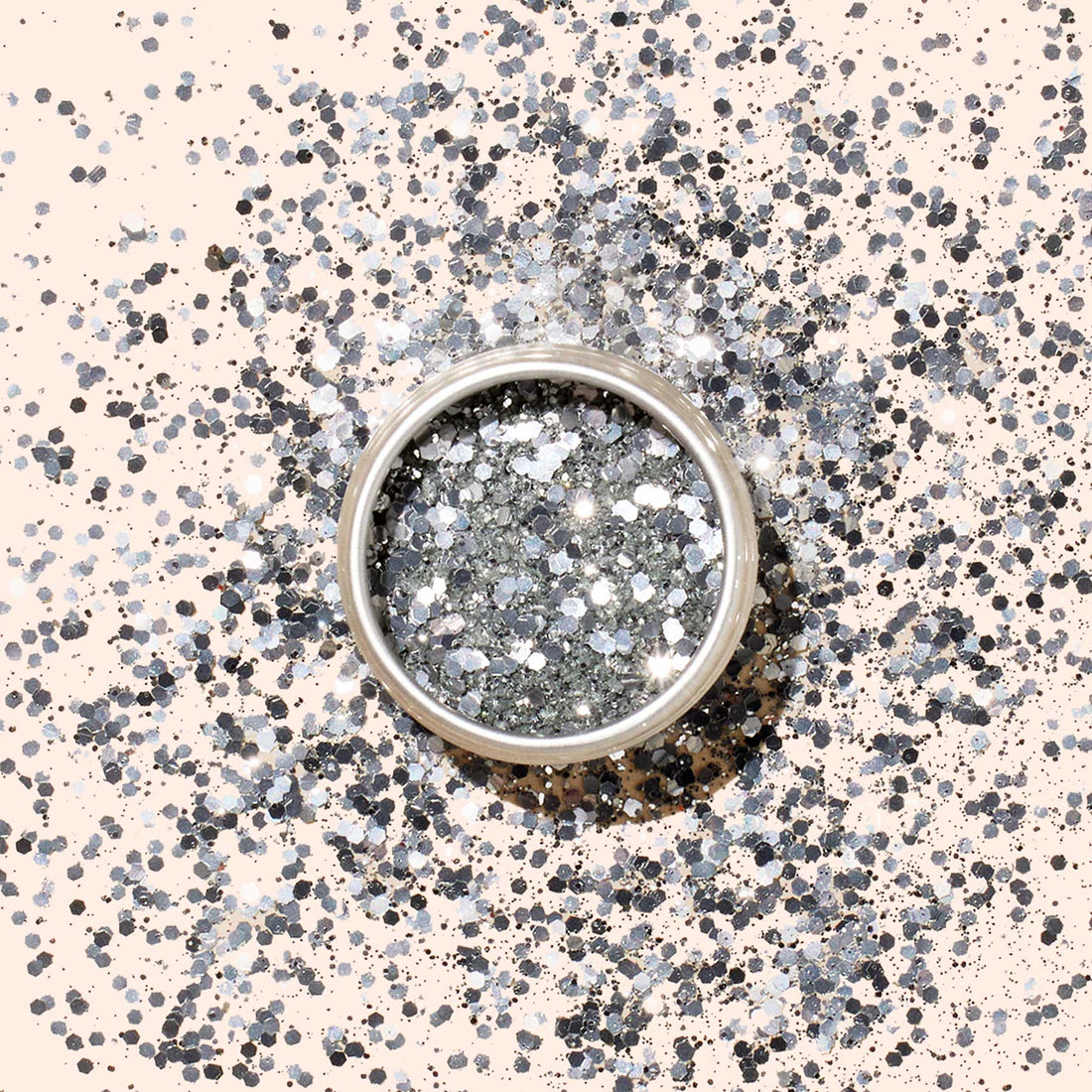 A round container of Manucurist Silver Biodegradable Glitter rests on a beige surface, surrounded by scattered silver sparkles—ideal for eco-friendly nail polish or festive silver nail art.