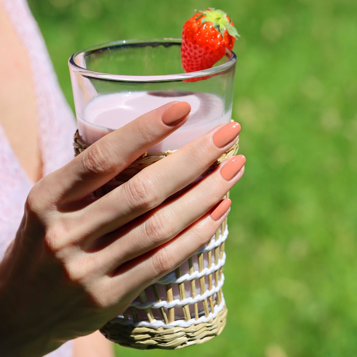 A person with Manucurist Peach nail polish holds a glass of pink strawberry milk topped with a fresh strawberry. The glass features a woven sleeve, set against a grassy outdoor backdrop.